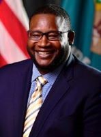 Photo of Director Cerron Cade smiling against the American and Delaware flags in the background.